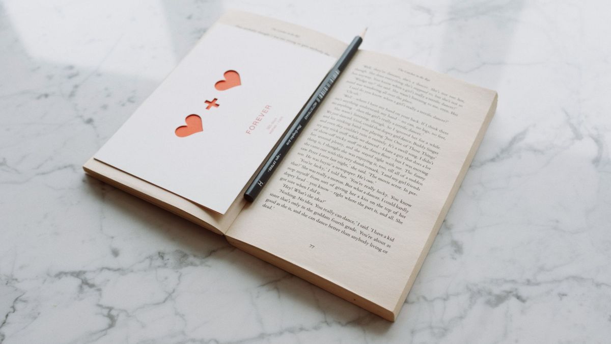 From above of opened novel book with pencil and romantic gift postcard composed on shiny white marble surface