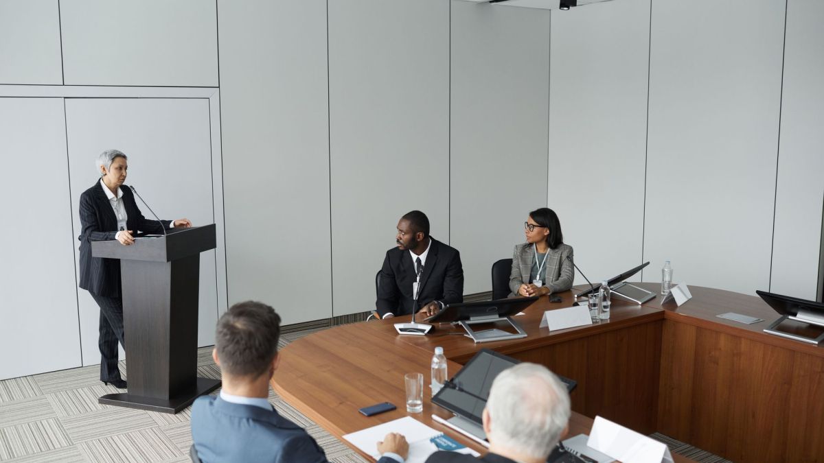 Diverse group of professionals in a formal meeting setting with a speaker at the podium.