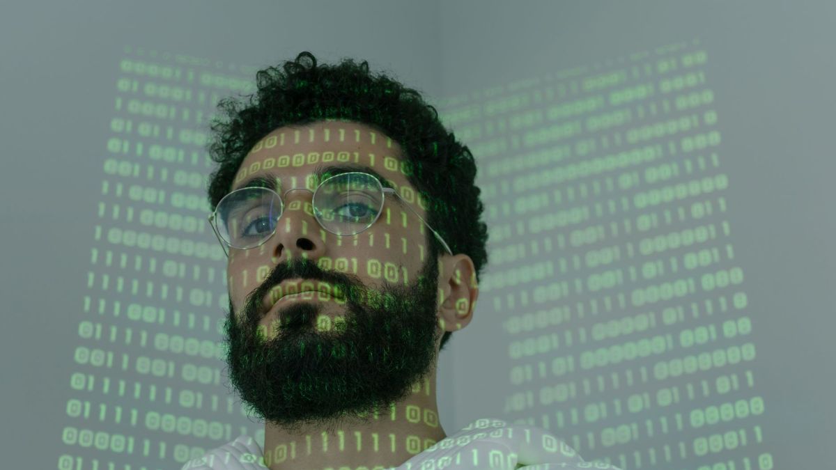 A man with a beard and eyeglasses looking at the camera, illuminated by binary code projection.