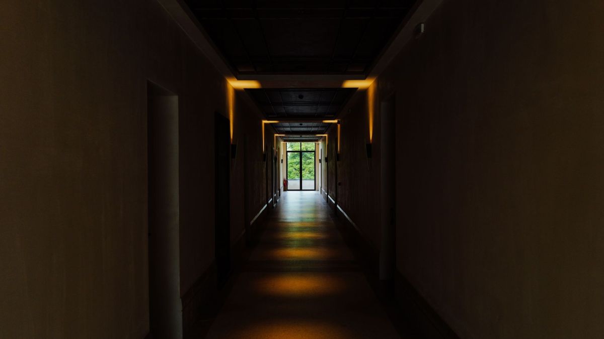 A shadowy indoor hallway with lights leading to a bright exit, creating a sense of mystery.