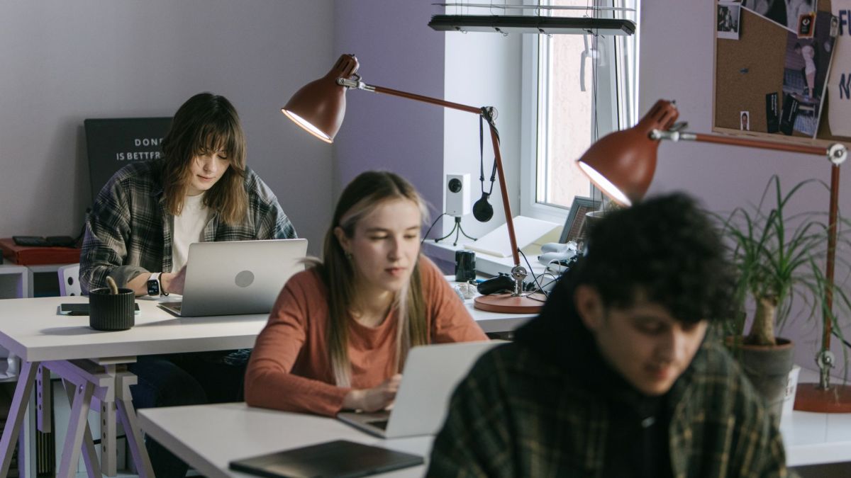 Three young professionals working at desks in a modern office setting with laptops and desk lamps.
