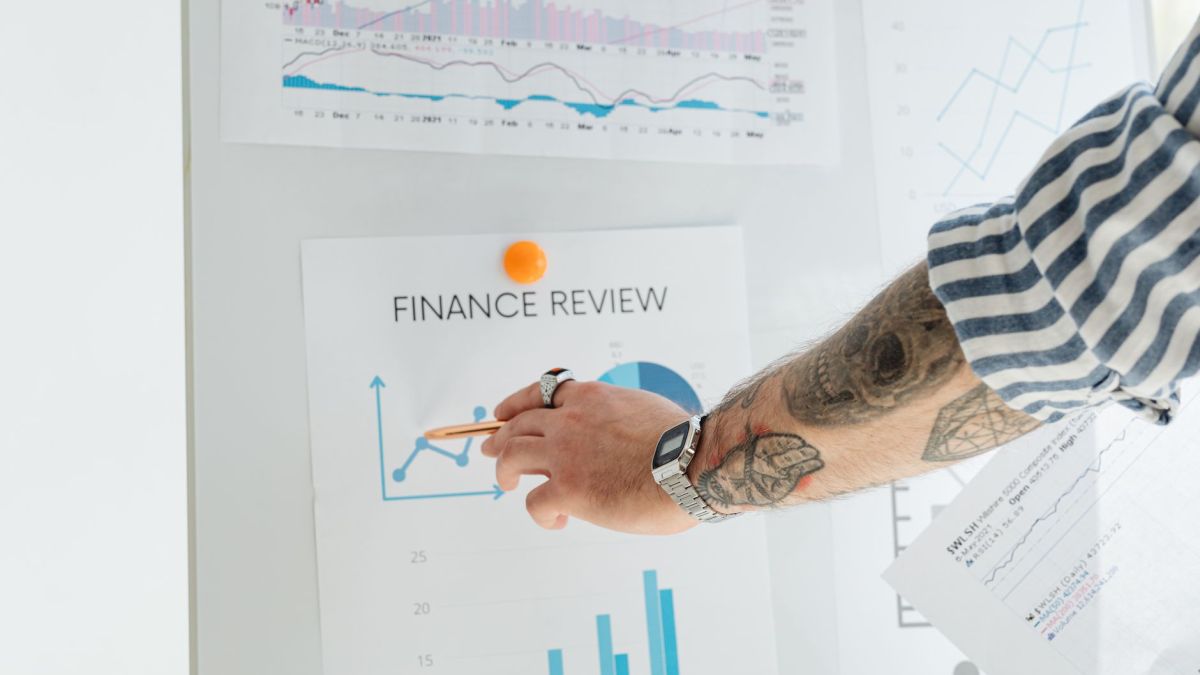 A person with tattooed arms presenting financial data using charts and graphs on a whiteboard.