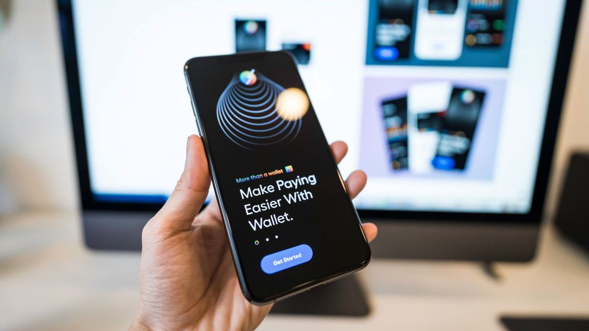 Hand holding smartphone displaying digital wallet app interface, blurred monitor in background.