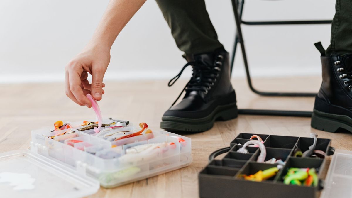 A person organizes fishing lures in plastic containers indoors, wearing black boots.