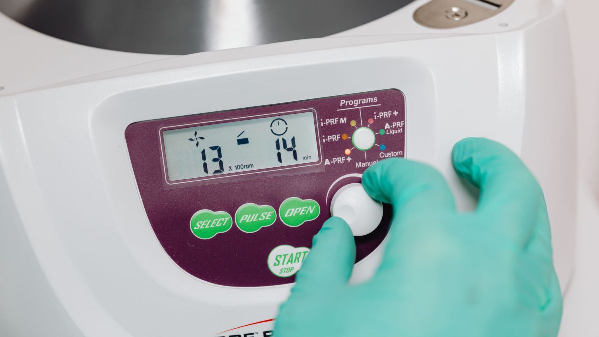 A gloved hand adjusts a control knob on a medical centrifuge device in a clinical setting.