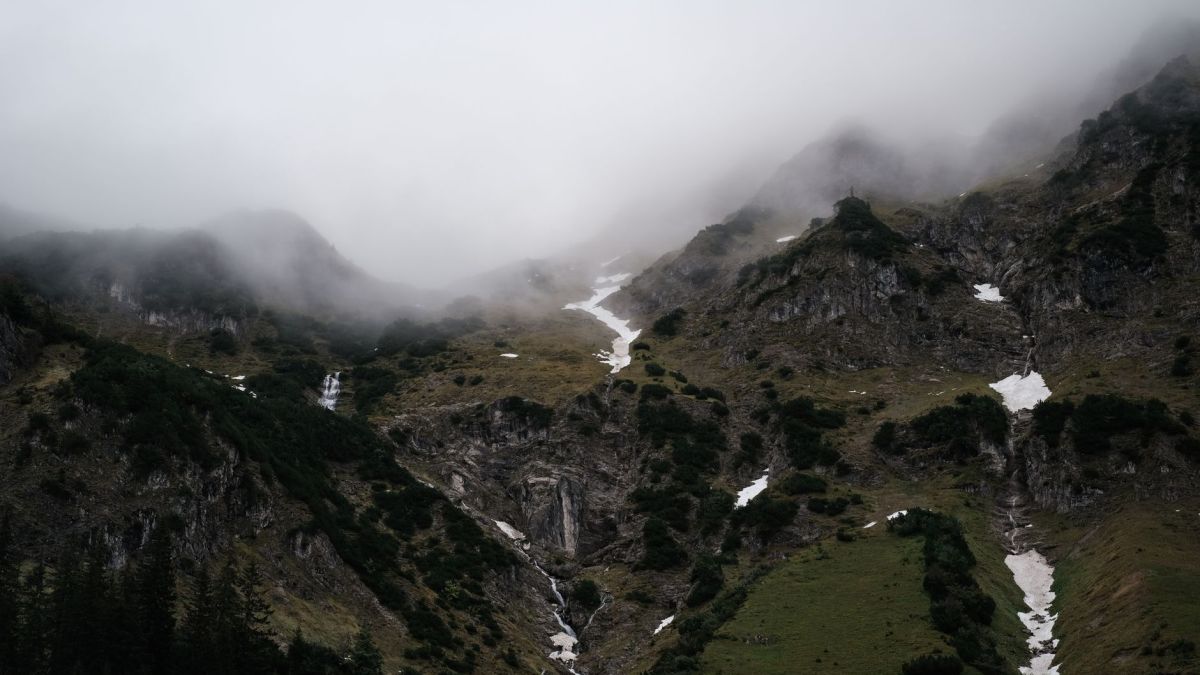 A scenic view of misty mountains with patches of snow and green foliage under cloudy skies.