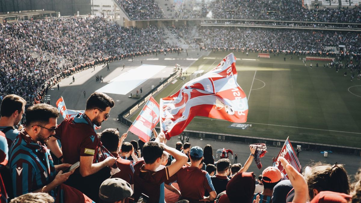 Vibrant crowd with flags cheering at a football stadium during the day.
