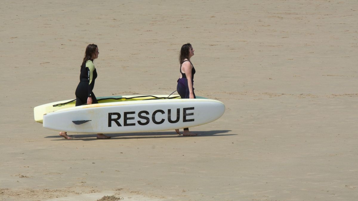 Two women in wetsuits walk on sandy beach carrying a rescue board under the summer sun.