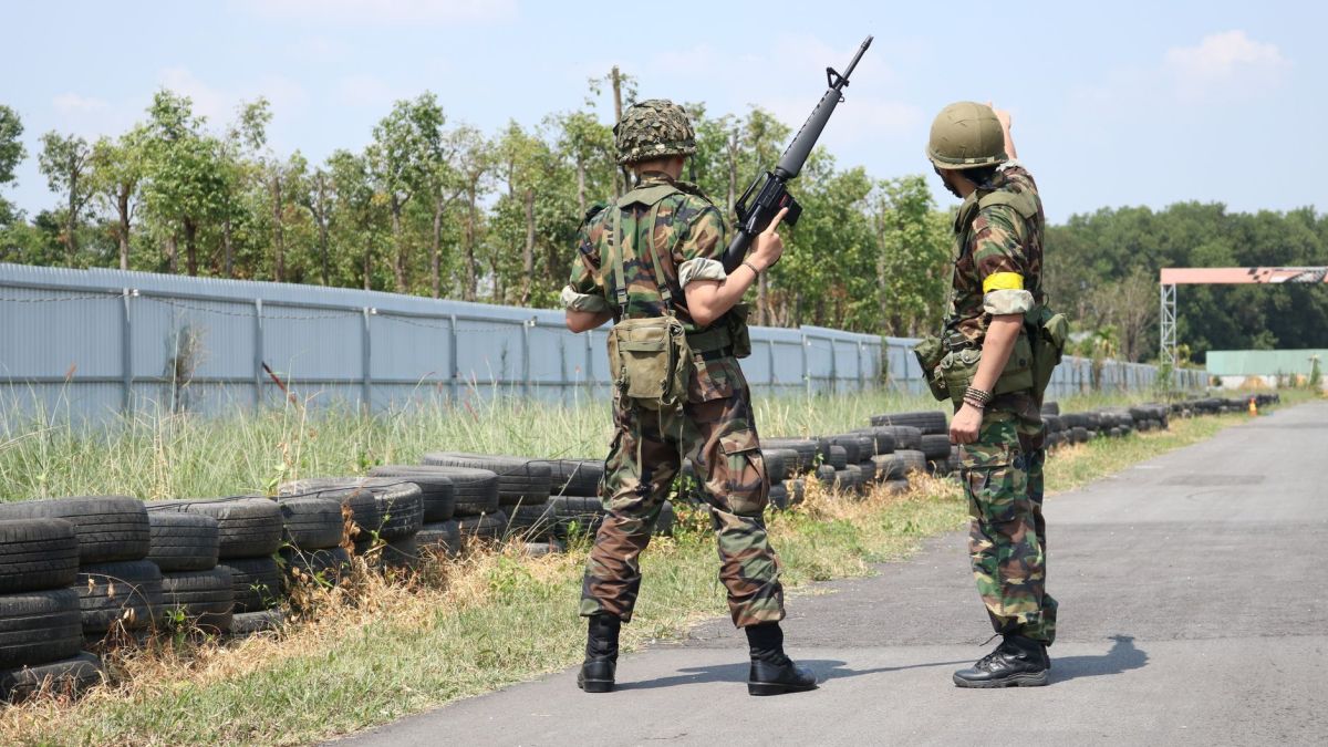 Two soldiers in camouflage uniforms with weapons stand outside on a training field.