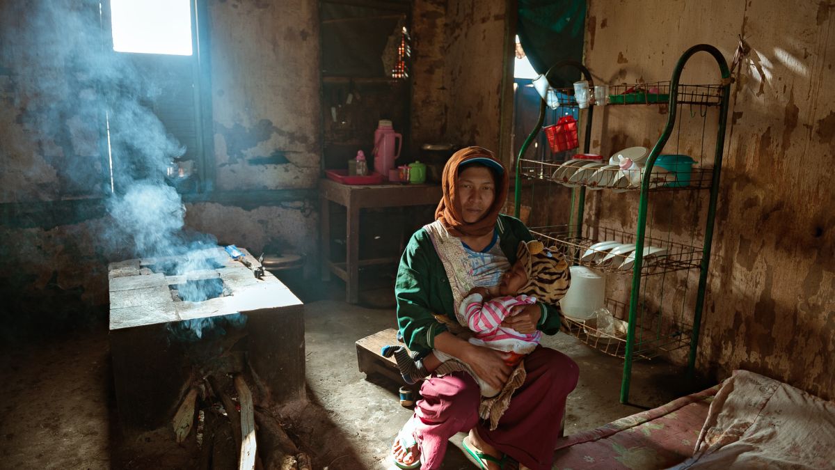 A mother holding her baby in a rustic, light-filled kitchen, with smoke rising from a stove.