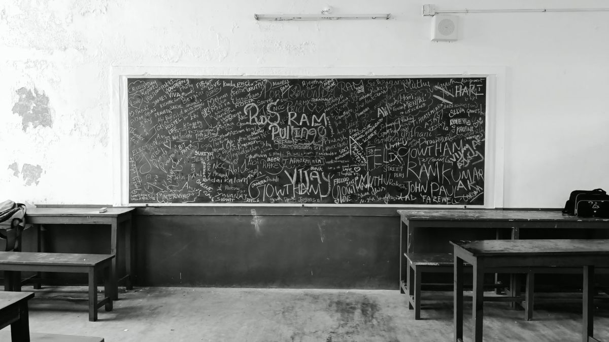 A nostalgic black and white photo of an empty classroom in Madurai, India, featuring a graffiti-covered chalkboard.