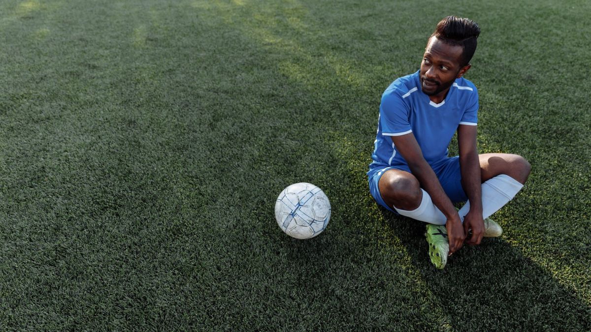 A soccer player in a blue jersey sits on the grass field during the day.