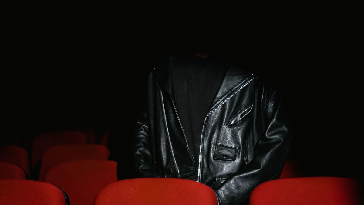 Individual wearing black leather jacket sits among red seats in a dimly lit cinema.