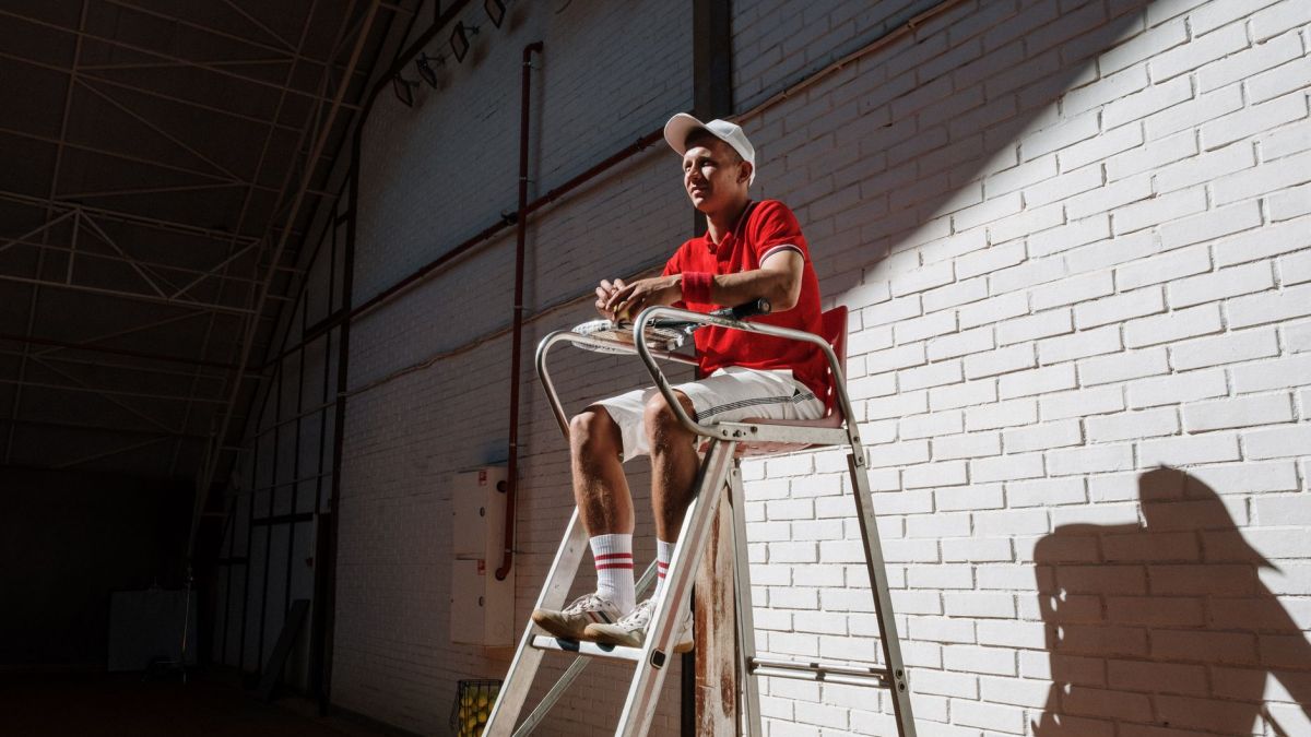 Tennis umpire sitting on a high chair against a white brick wall, creating dynamic shadows indoors.