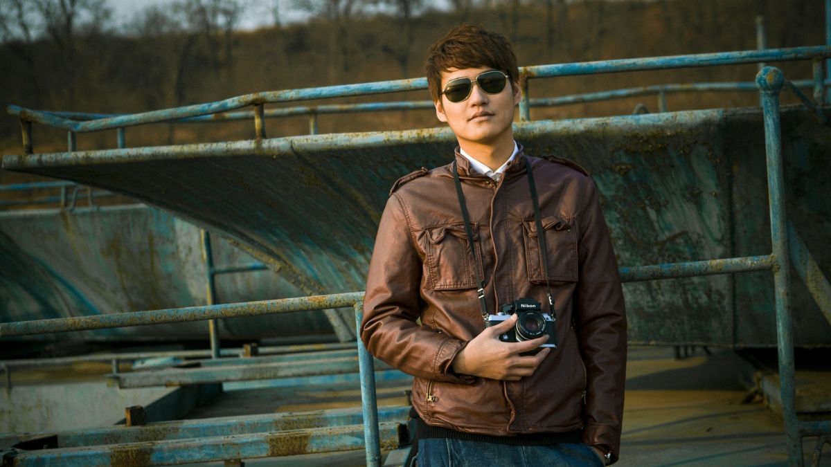 A man in sunglasses and jacket holds a camera in an outdoor industrial setting.