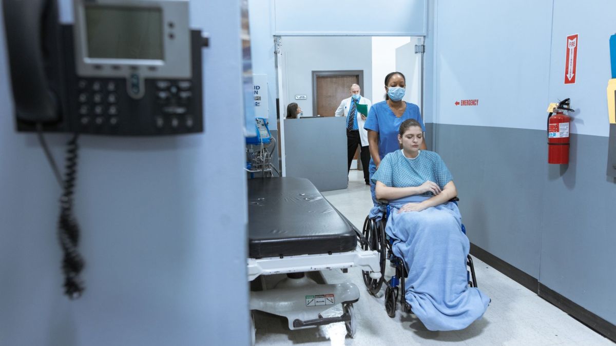 A nurse helps a patient in a wheelchair down a hospital corridor, reflecting care and medical professionalism.