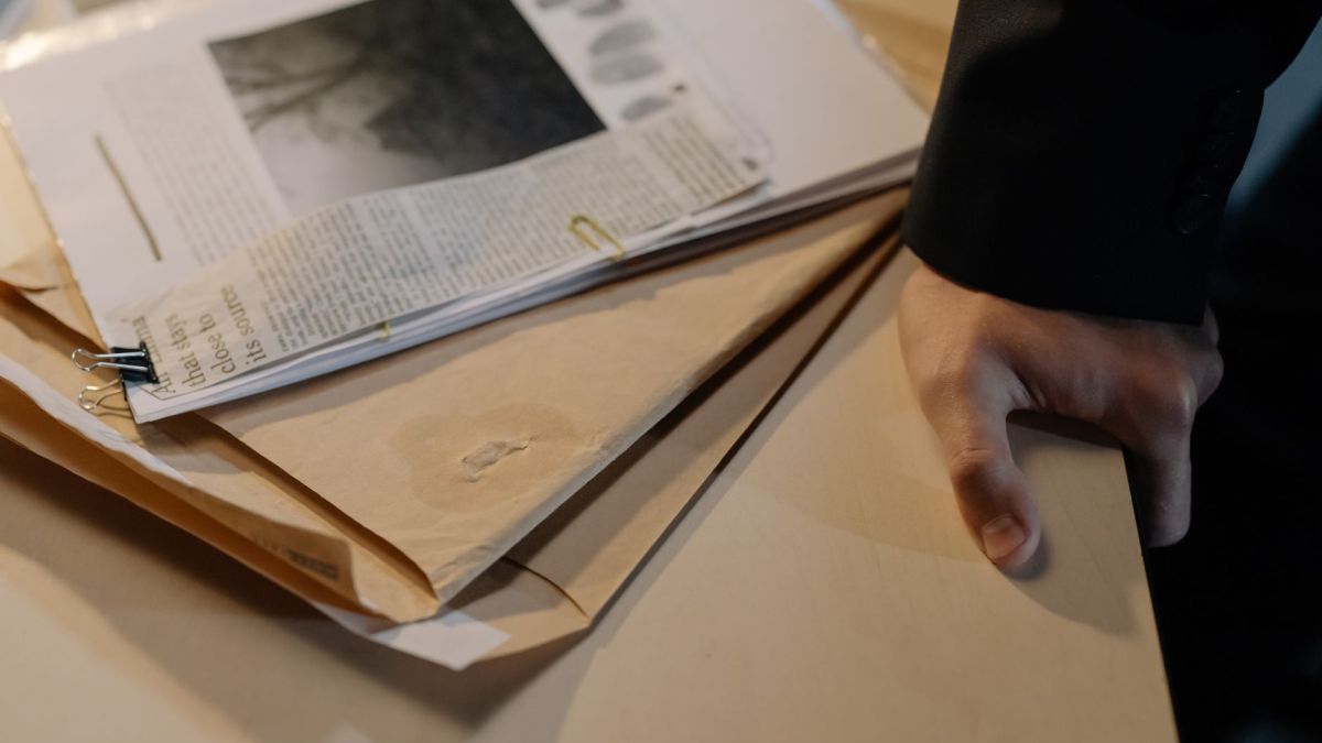 Close-up of a hand resting on a desk near a stack of investigative documents and newspaper clippings.