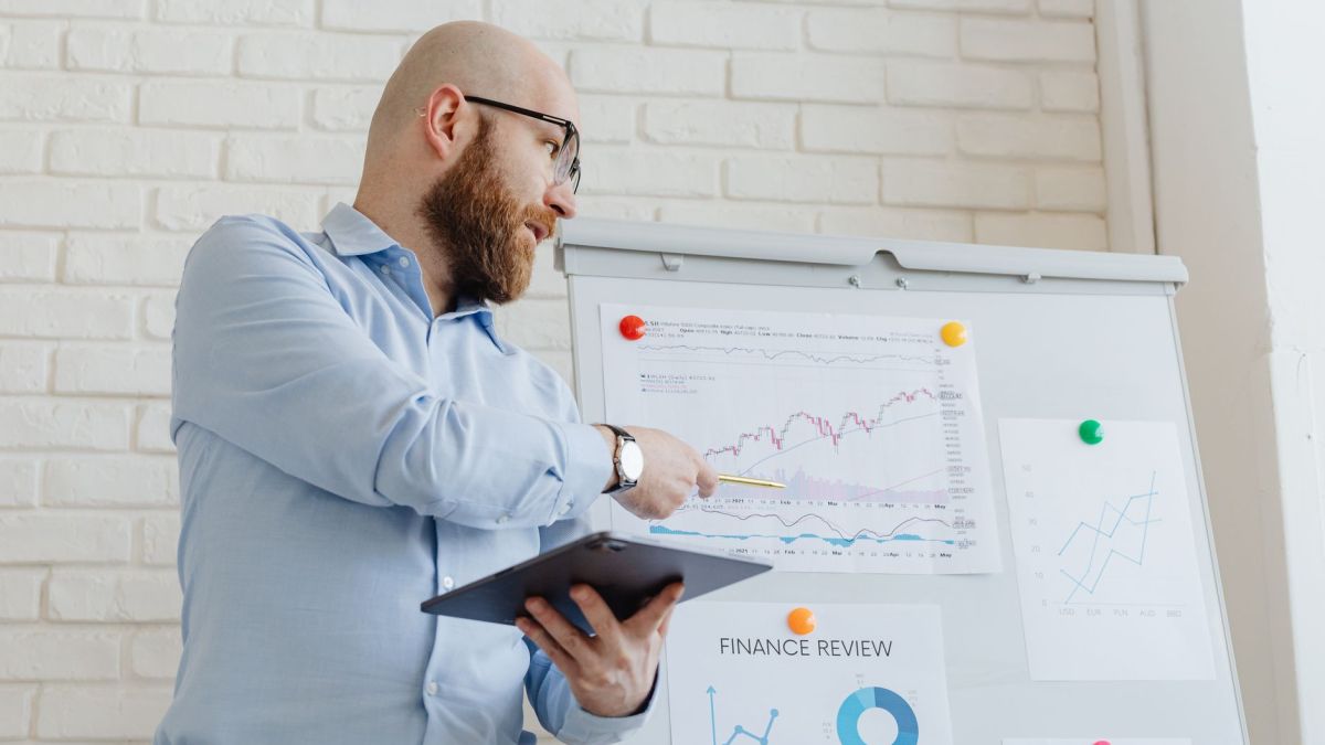 Man giving a business presentation with financial graphs on a whiteboard using a tablet for explanation.