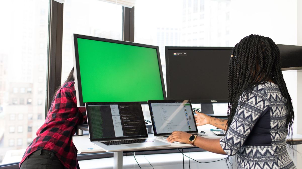 Two women working on laptops and monitors in a bright office setting, focused on technology and teamwork.