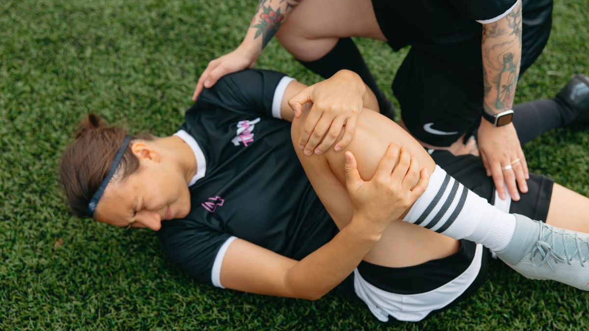 A female soccer player lying on grass, holding her injured knee, supported by a teammate.