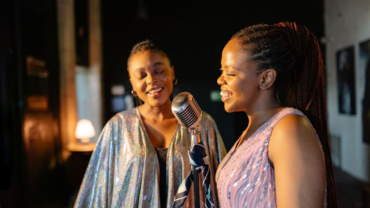 Two women performing indoors, sharing a vintage microphone in joyful harmony.