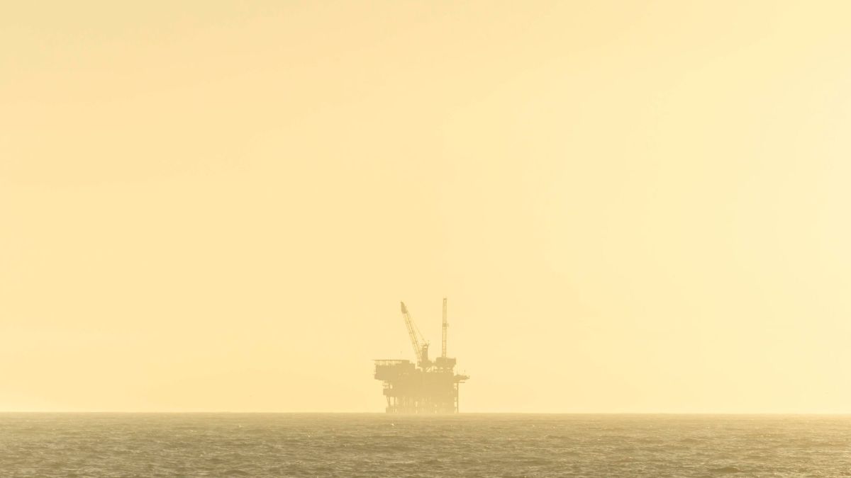 Distant view of an offshore oil platform against a golden sky at sunset over a calm sea.