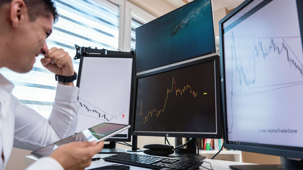 Trader in white shirt analyzing stock charts on multiple monitors during daytime in an office setting.