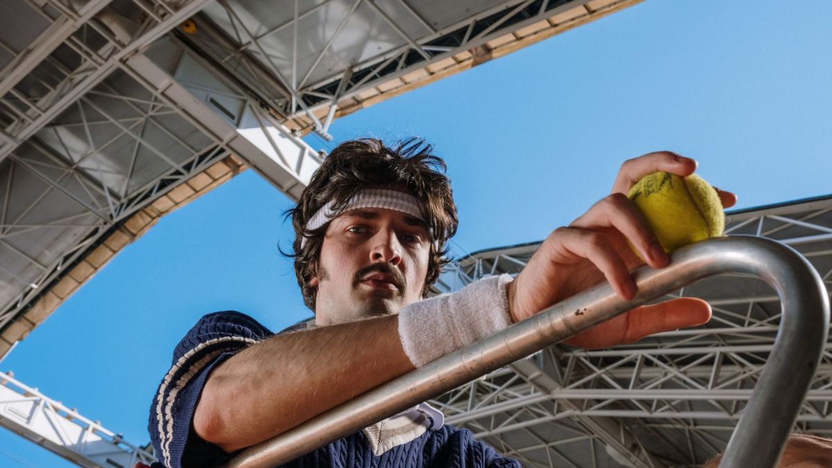 Low angle view of a tennis player holding a ball beneath a stadium roof. Bright, clear sky.