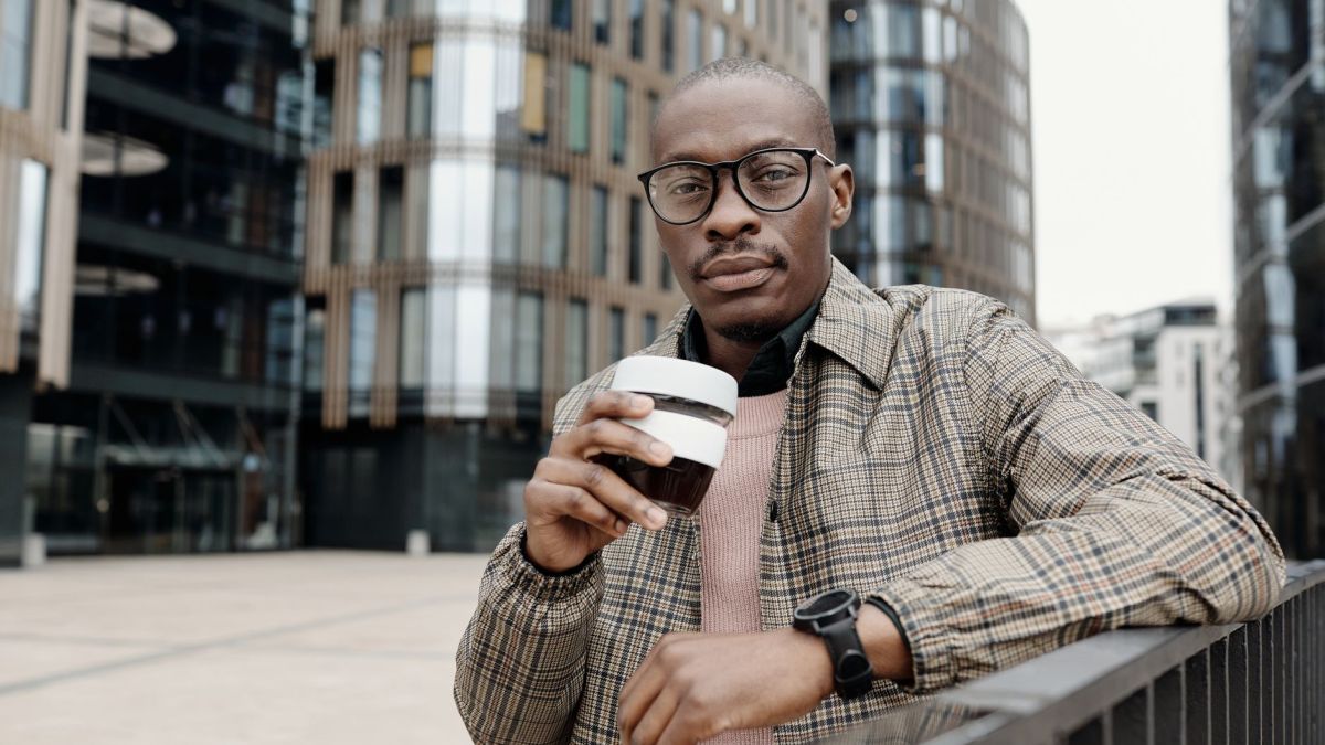 Confident man in glasses holding a coffee cup in a modern urban area, exuding style and sophistication.