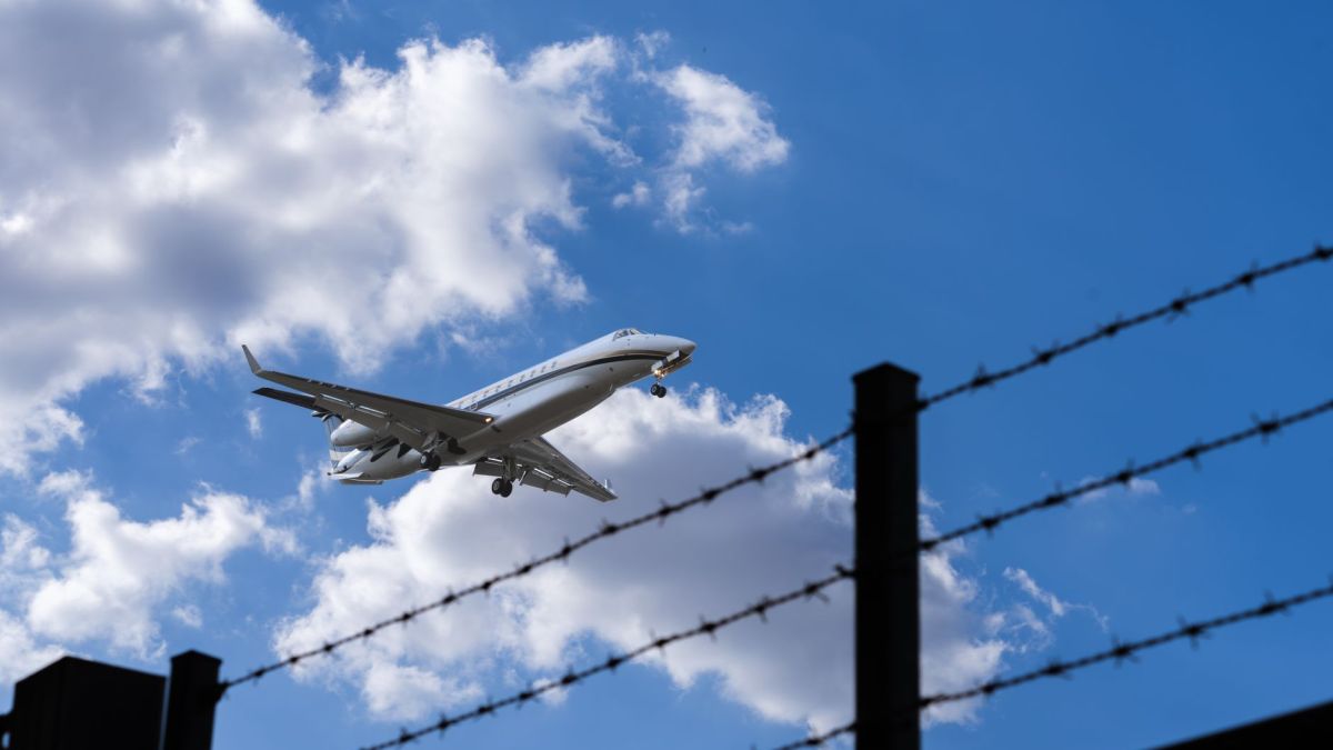 Jet approaching for landing, silhouetted against a bright blue sky with clouds and barbed wire in foreground.
