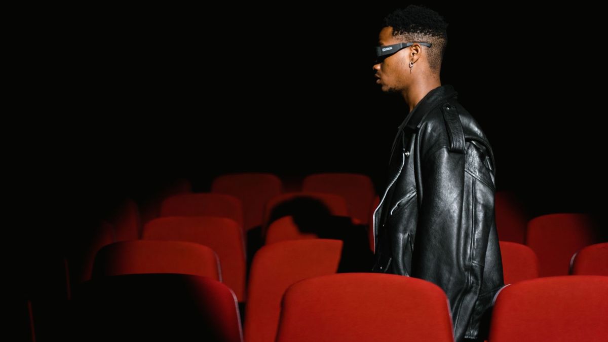 A man wearing glasses walks through a dimly lit movie theater with red seats.