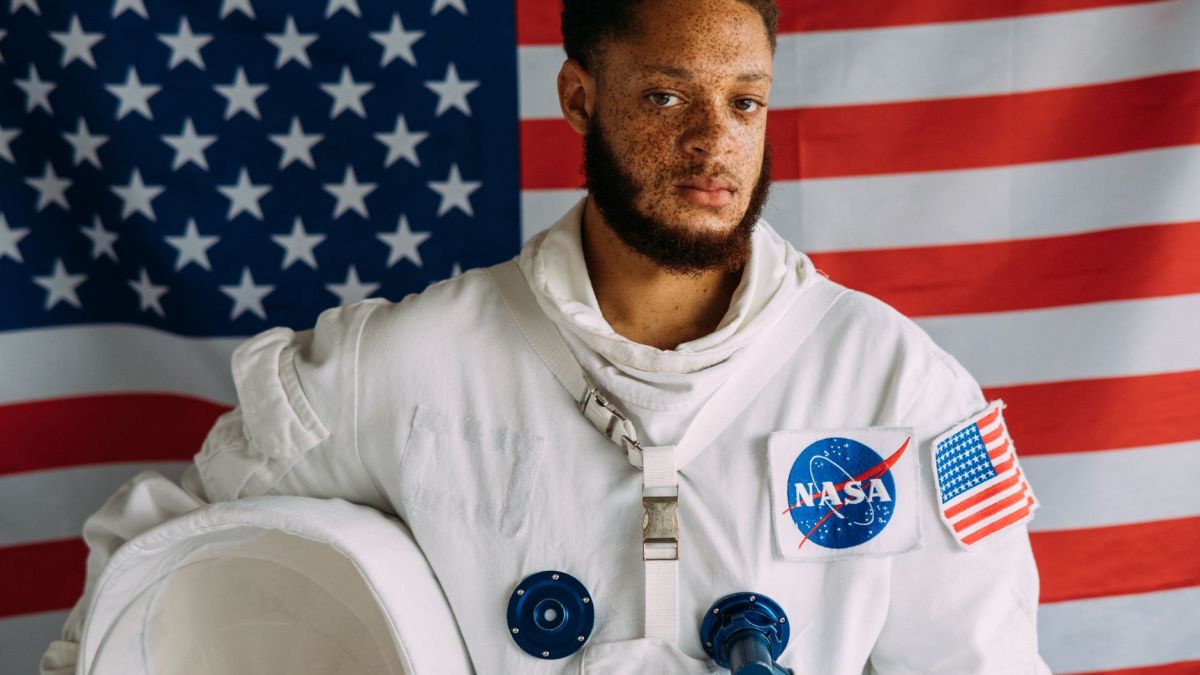 Portrait of an African American man in a NASA astronaut suit against an American flag backdrop.