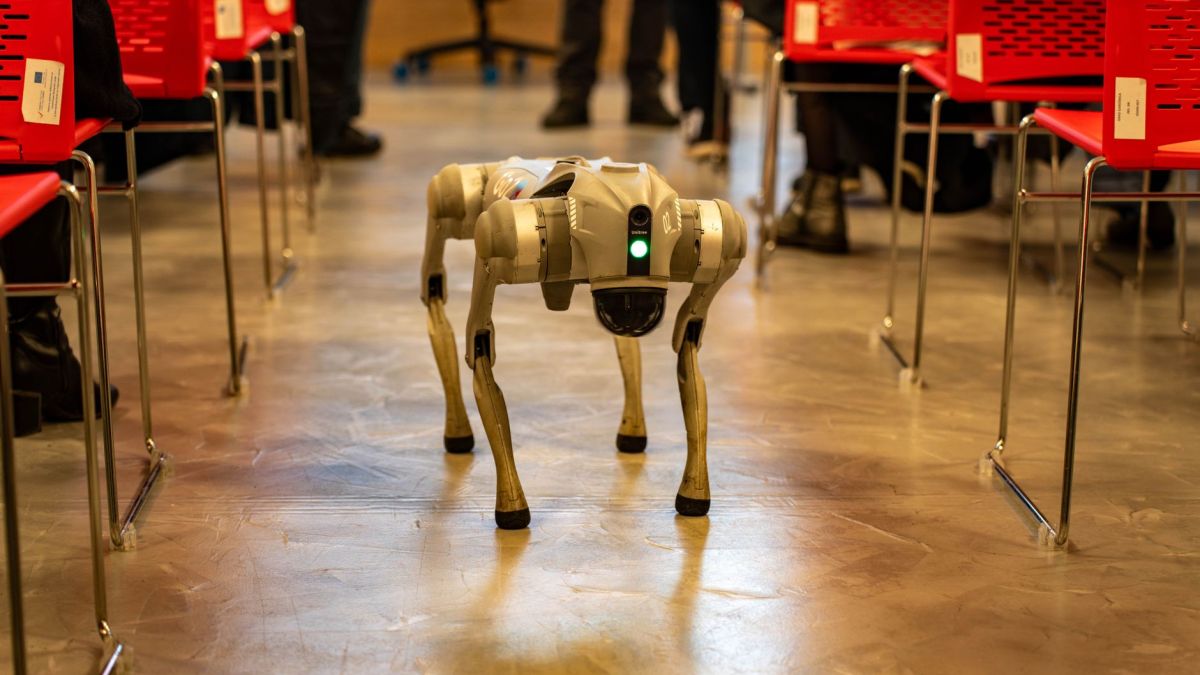 A robotic dog navigates an indoor setting amidst red chairs, showcasing technology in modern environments.