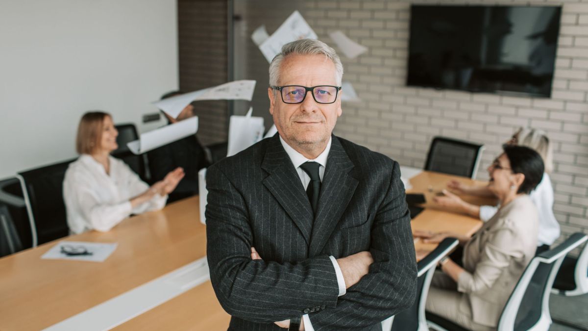 Business executive standing confidently in meeting room with team engaged in discussion behind.