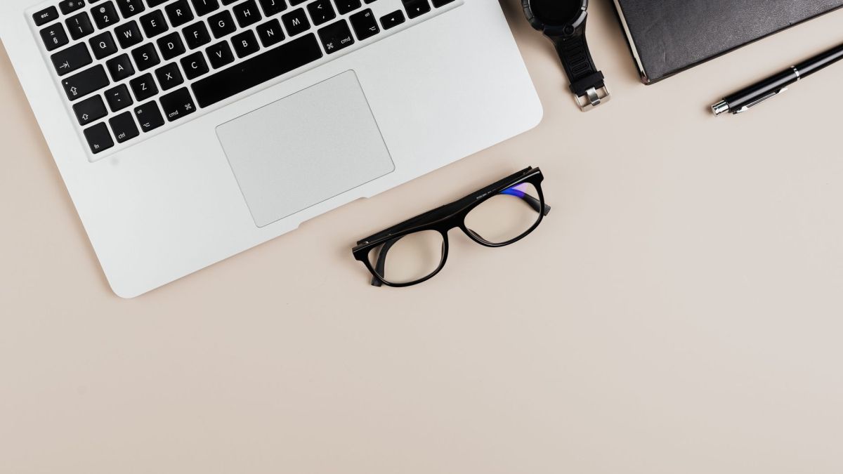 Modern workspace flat lay with laptop, notebook, watch, and glasses on a beige background.