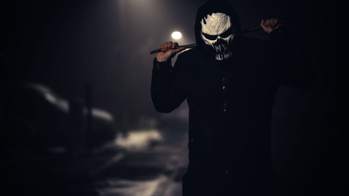 A spooky masked figure holding a weapon in a dimly lit street at night.