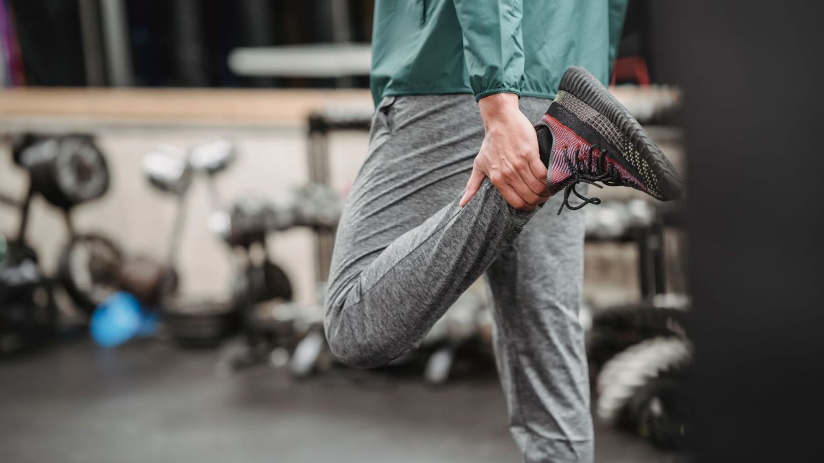Crop anonymous male in sportswear warming up while stretching legs on blurred background of sport club with gym equipment