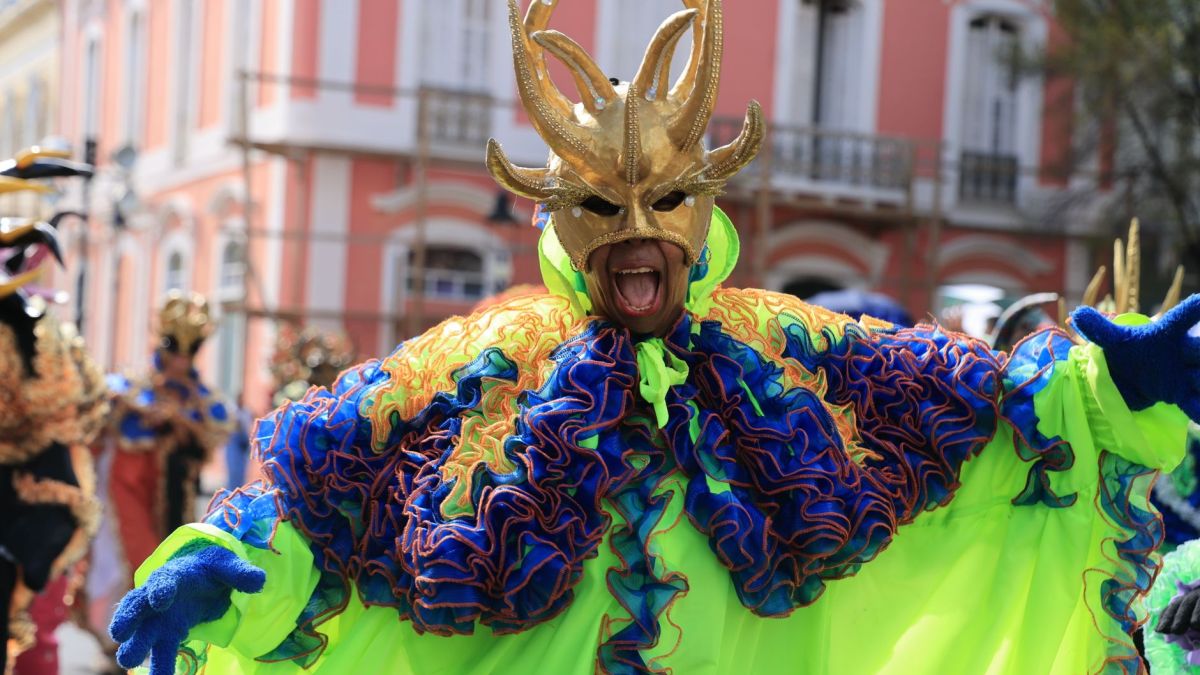 A performer in colorful costume and mask at a lively outdoor carnival parade.