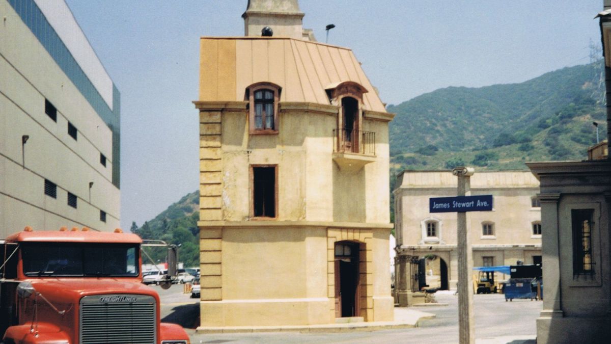 A rustic building on a film studio lot with a parked truck near a mountain background.