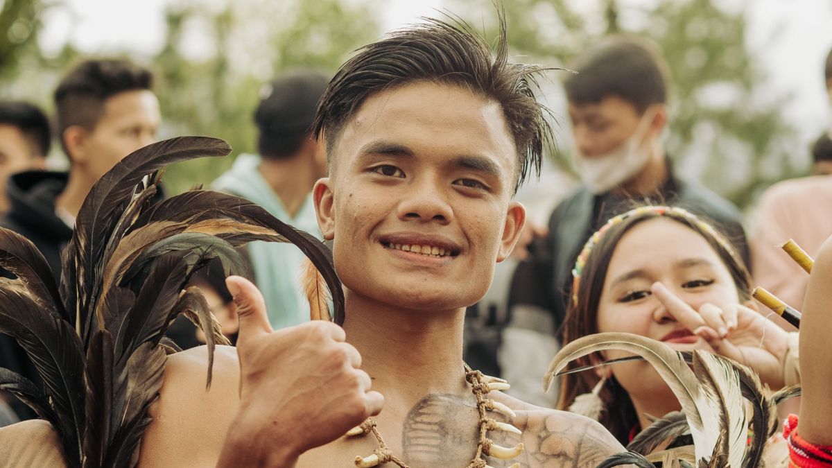 Cheerful man giving thumbs up in tribal attire with feathers at outdoor event.