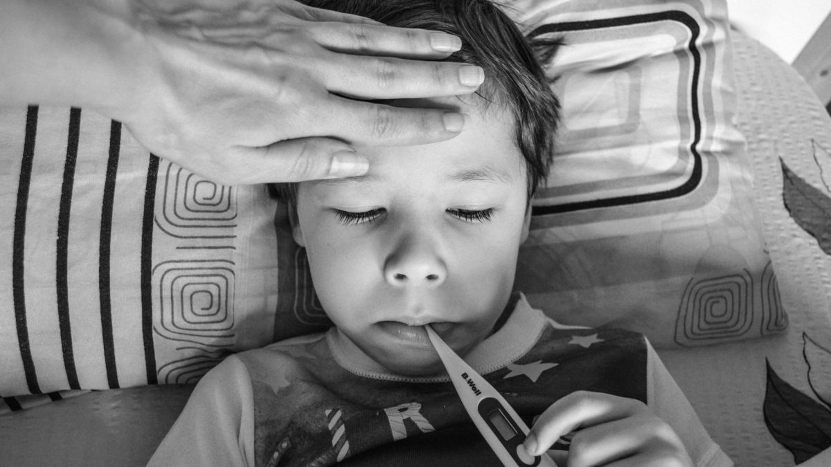 Black and white from above of unrecognizable person touching forehead of unemotional child in casual clothing holding electronic thermometer in mouth