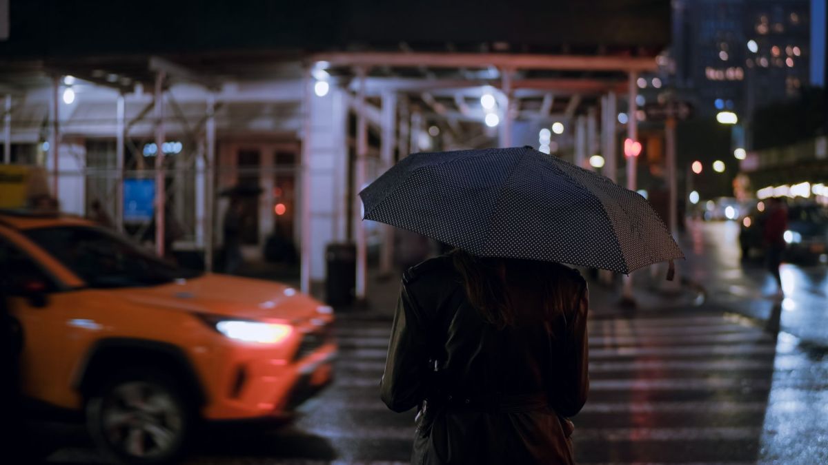 A person holding an umbrella crosses a wet city street at night under urban lights.