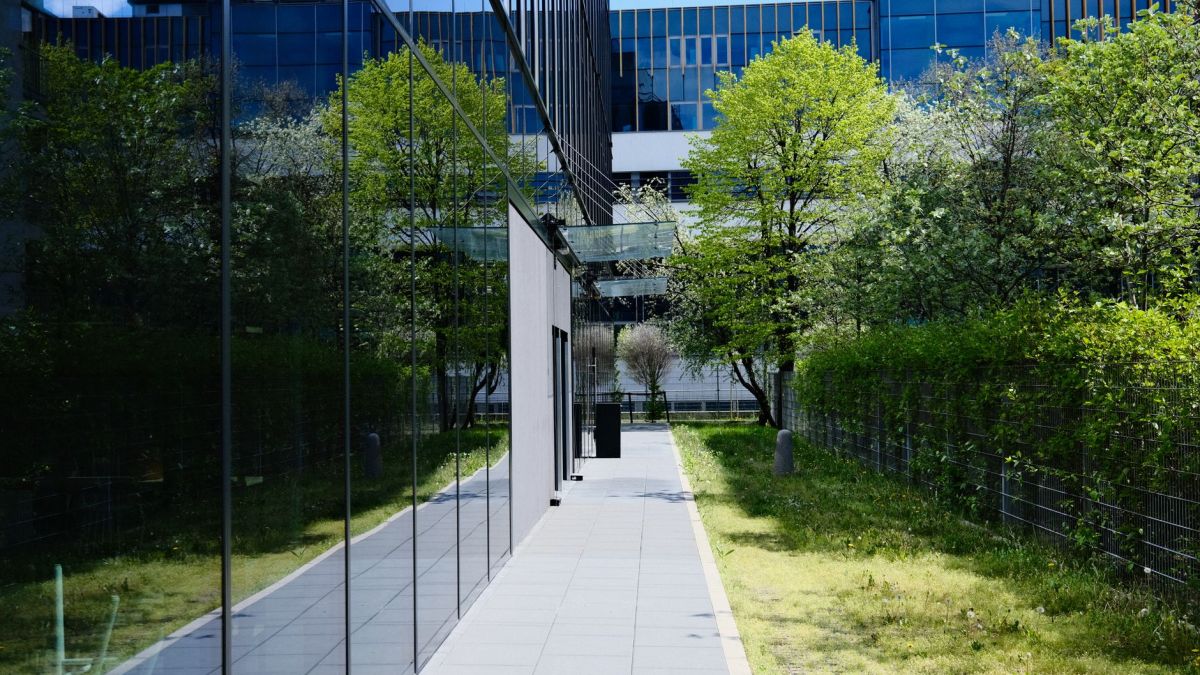 Modern glass building with pedestrian walkway and greenery, reflecting urban architecture.