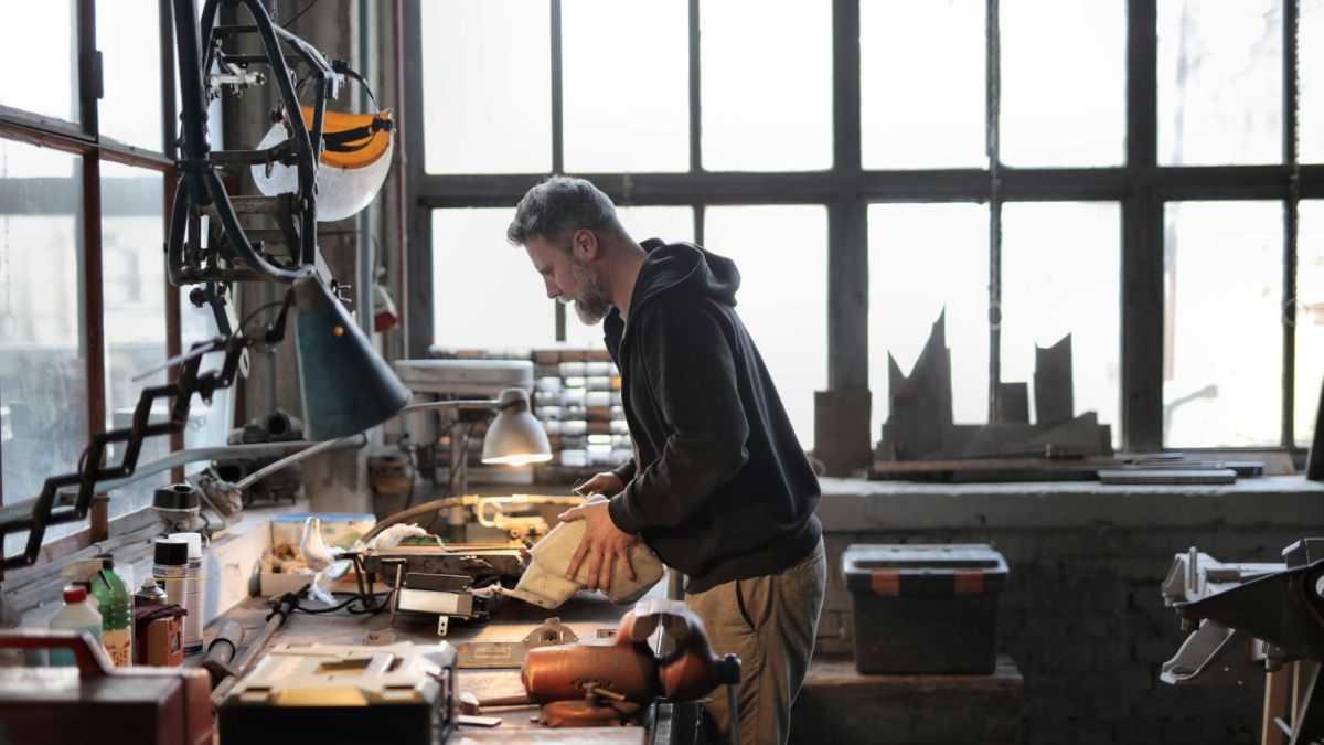 Side view of male workshop owner standing near messy workbench and fixing metal details while using instruments during work