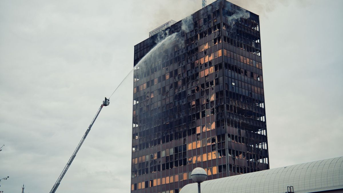 Firefighter using hose to extinguish fire in a tall building under cloudy sky.