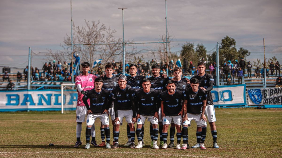 A soccer team poses together on an outdoor field before a match, with spectators in the background.