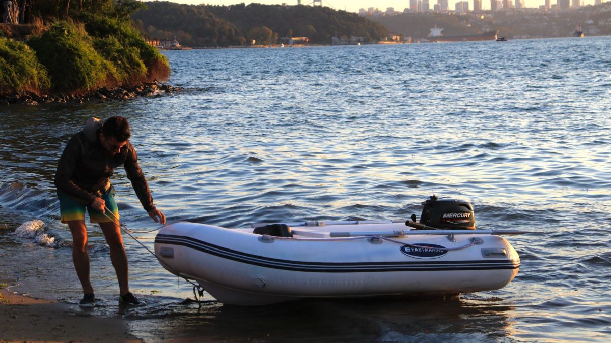 A man pulls an inflatable boat onto a sandy shore at sunset with a city skyline in the background.