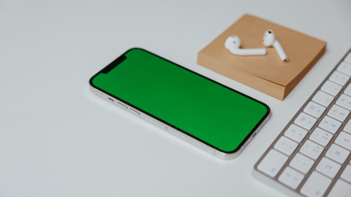 A white smartphone with a green screen next to wireless earphones and a keyboard on a white surface.