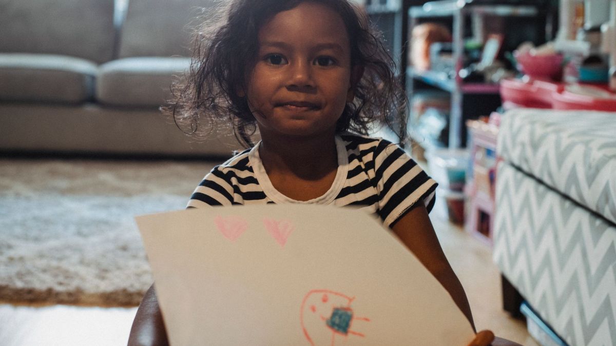 Cute ethnic girl in casual shirt demonstrating paper sheet with drawing while sitting in living room and looking at camera