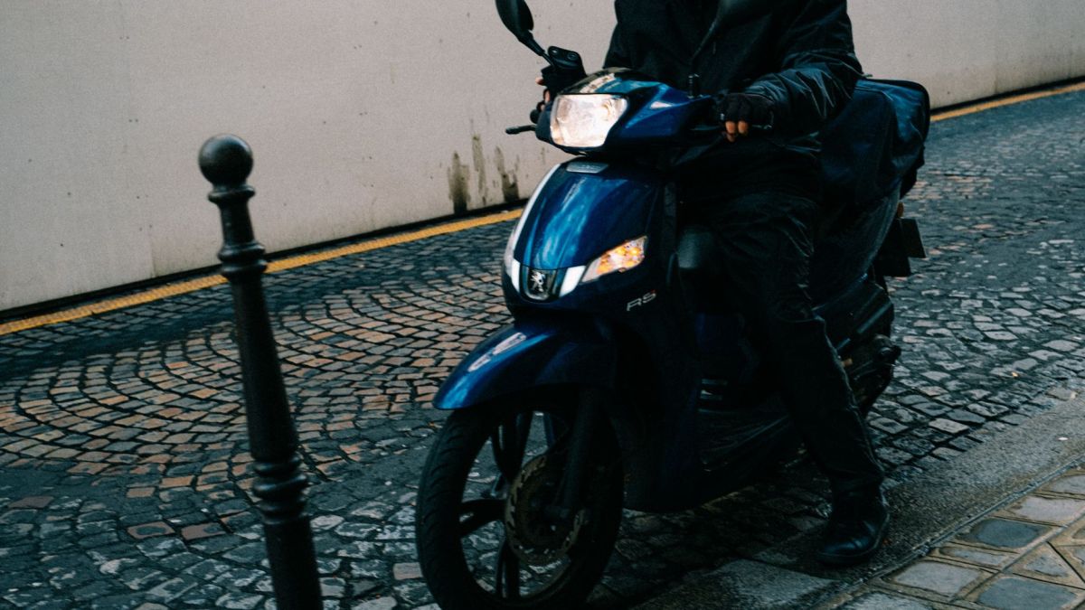 A motorcyclist wearing a helmet and face mask rides a scooter on a cobblestone street.